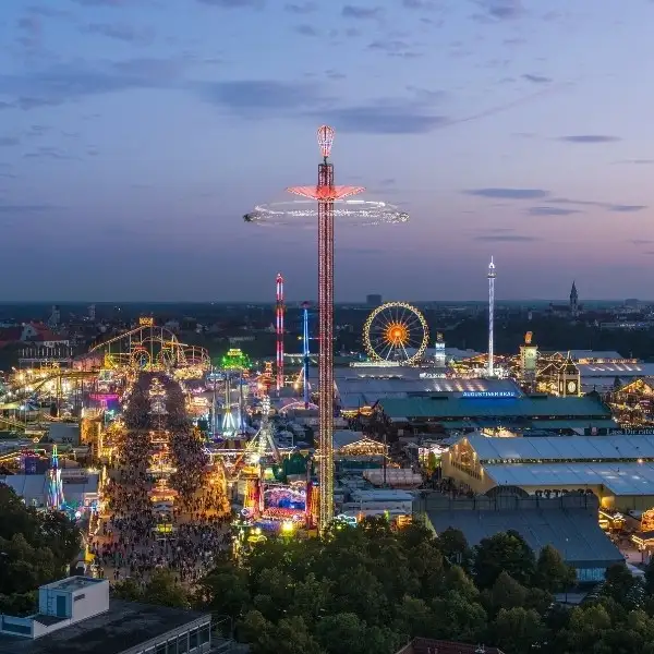 Nighttime aerial view of the Oktoberfest fairgrounds lit up with colorful lights, ferris wheels, rides, and bustling crowds.